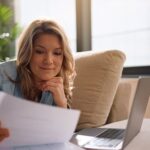 Frau sitzt entspannt auf dem Sofa, liest ein Schreiben und hat den Laptop mit ihren Finanzunterlagen vor sich