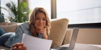 Frau sitzt entspannt auf dem Sofa, liest ein Schreiben und hat den Laptop mit ihren Finanzunterlagen vor sich