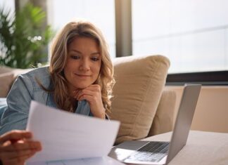 Frau sitzt entspannt auf dem Sofa, liest ein Schreiben und hat den Laptop mit ihren Finanzunterlagen vor sich