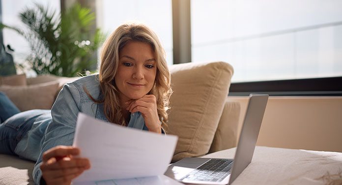 Frau sitzt entspannt auf dem Sofa, liest ein Schreiben und hat den Laptop mit ihren Finanzunterlagen vor sich