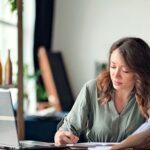 Kleinunternehmerregelung 2026: Was sie wirklich bedeutet und wann sie sich lohnt Frau sitzt im Homeoffice am Schreibtisch, prüft Unterlagen und macht sich Notizen neben ihrem Laptop