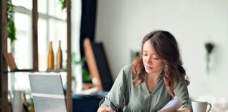 Frau sitzt im Homeoffice am Schreibtisch, prüft Unterlagen und macht sich Notizen neben ihrem Laptop