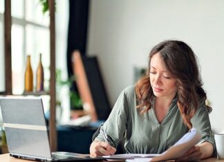 Frau sitzt im Homeoffice am Schreibtisch, prüft Unterlagen und macht sich Notizen neben ihrem Laptop
