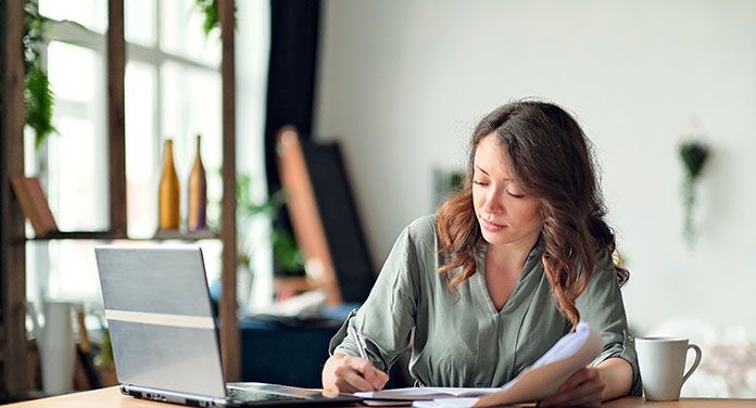 Frau sitzt im Homeoffice am Schreibtisch, prüft Unterlagen und macht sich Notizen neben ihrem Laptop