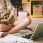 Frau sitzt auf dem Boden, notiert Zahlen in einem Notizbuch, daneben steht ein aufgeklappter Laptop